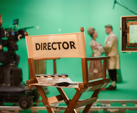 Film Studio set focus on empty director's chair. In the background professional crew shooting historic movie, cameraman on railway trolley shooting green screen scene with actors for history movie