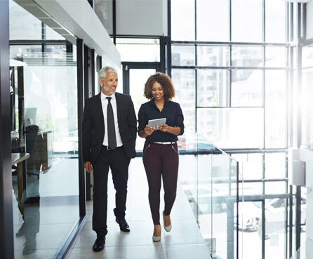 Two business professionals—a man and a woman—walking through a modern office corridor, smiling and reviewing information on a tablet.