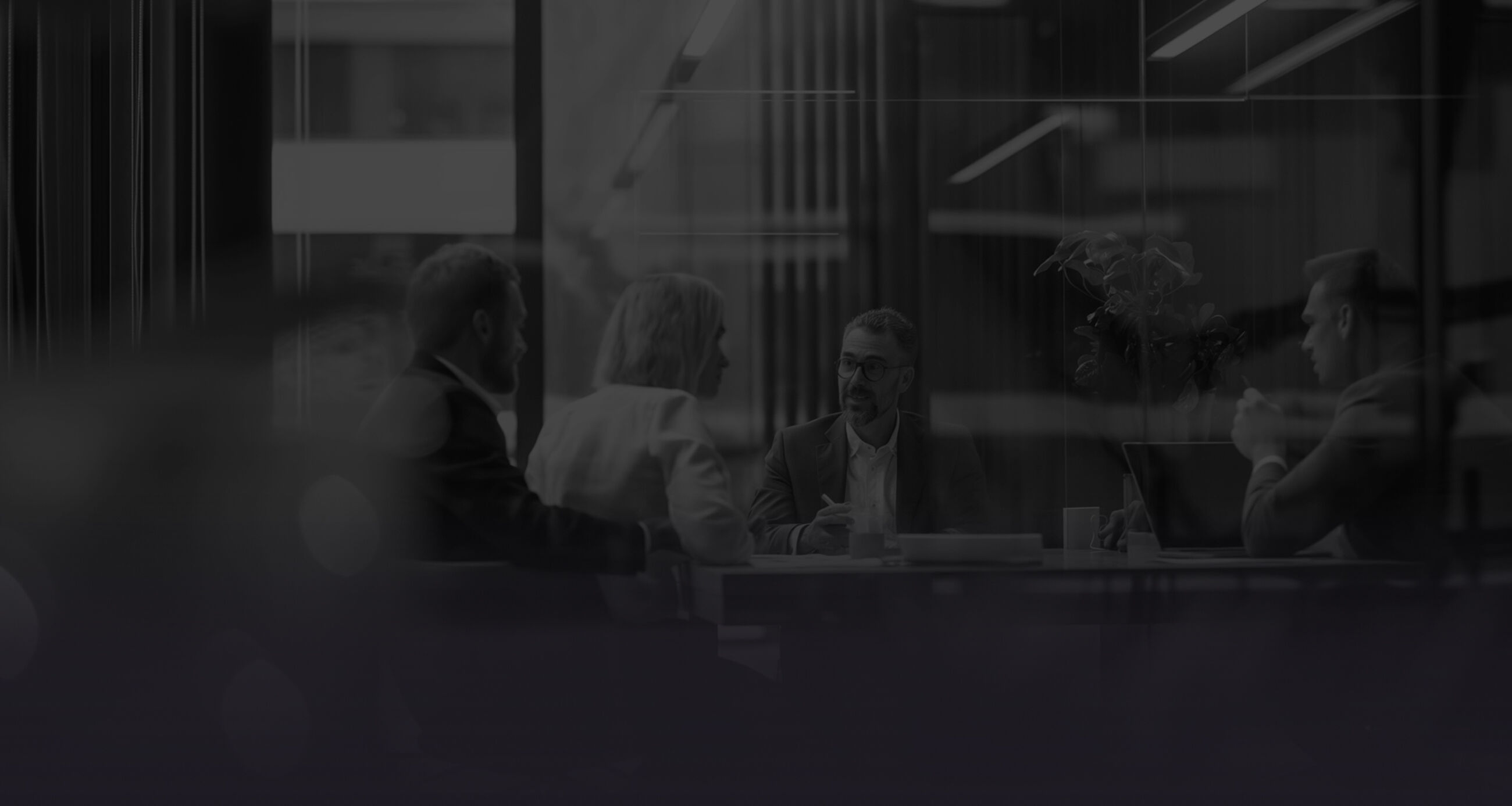 Four business professionals sitting around a meeting table in a modern glass-walled office, engaged in conversation.
