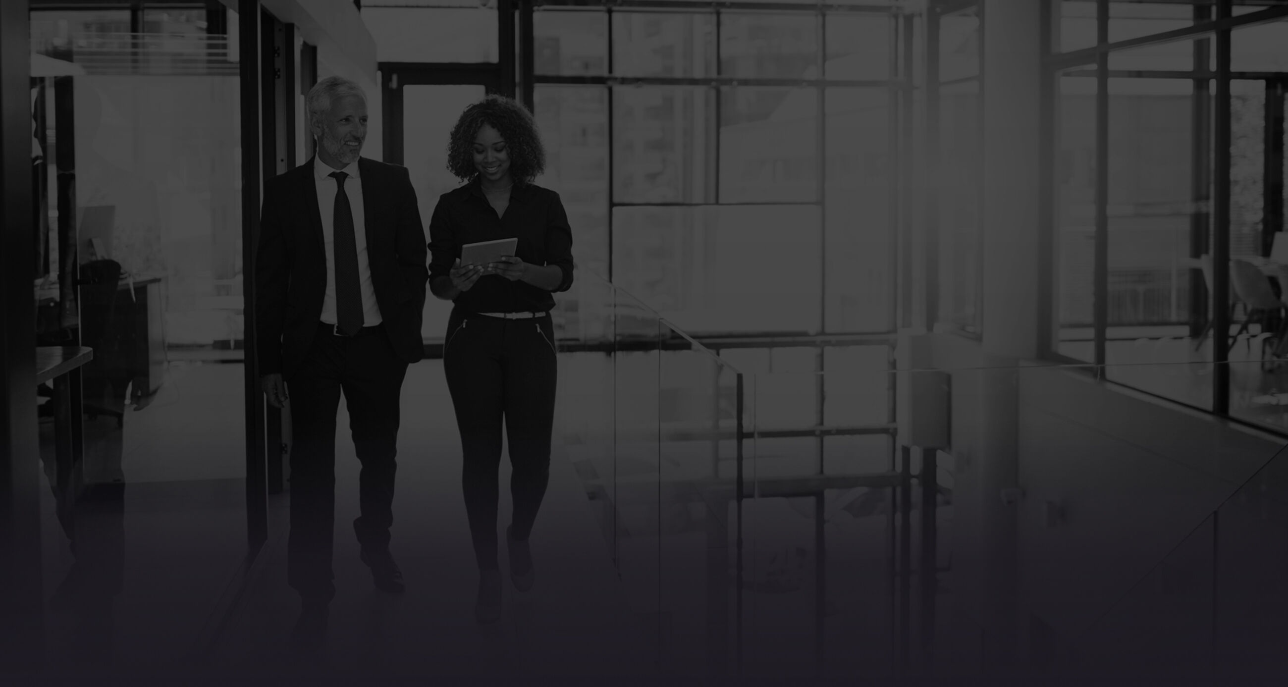 Two business professionals—a man and a woman—walking through a modern office corridor, smiling and reviewing information on a tablet.